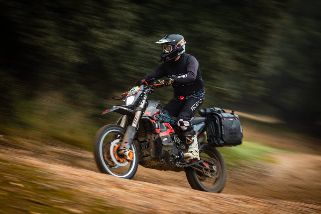 Motorista en una KTM 890 Rally equipada con maletas Sysbag WP, rodando con velocidad por un camino de tierra en un entorno forestal